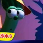 Larry the cucumber puppet wearing a yellow hat on a colorful VeggieTales stage, with a poster on the wall in the background.