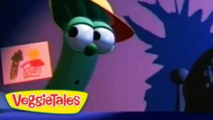 Larry the cucumber puppet wearing a yellow hat on a colorful VeggieTales stage, with a poster on the wall in the background.