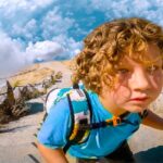 Young child wearing a teal shirt and backpack, on a rocky trail, studying a torn treasure map under a blue sky with clouds.