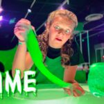 Woman wearing a green apron stretches neon green slime toward the camera in a bright, lab-like room.