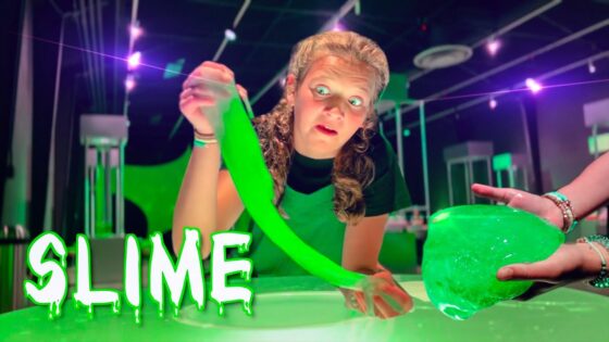 Woman wearing a green apron stretches neon green slime toward the camera in a bright, lab-like room.