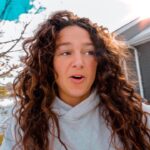 Close-up of a woman with long curly brown hair wearing a light gray hoodie, outdoors near a gray house with snow on the ground and a bright sun flare in the top-right corner.