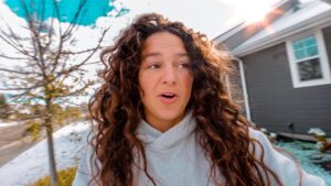 Close-up of a woman with long curly brown hair wearing a light gray hoodie, outdoors near a gray house with snow on the ground and a bright sun flare in the top-right corner.