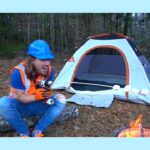 Man in a blue hard hat and orange safety vest sits by a small camping tent in a forest, holding a cordless drill.