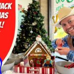 Smiling man in a cowboy hat and vest decorates a gingerbread house at a festive table, with a Christmas tree in the background.