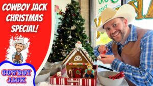Smiling man in a cowboy hat and vest decorates a gingerbread house at a festive table, with a Christmas tree in the background.