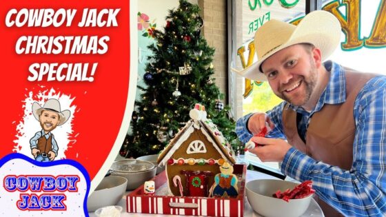 Smiling man in a cowboy hat and vest decorates a gingerbread house at a festive table, with a Christmas tree in the background.