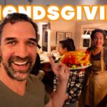 Selfie-style shot of friends at a kitchen party for Friendsgiving, a man in front smiles while presenting a plate of roasted turkey and sides to the group behind him.