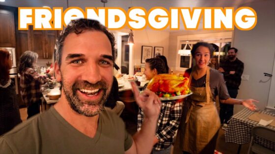 Smiling man in the foreground holds a plate with roasted turkey at a Friendsgiving gathering in a warm kitchen.