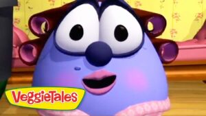 Blue, chubby VeggieTales character with large eyes, pink lips, and rosy cheeks smiles in a colorful room; VeggieTales logo bottom-left.