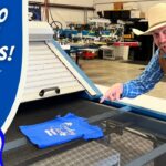 Man wearing a cowboy hat and vest demonstrates screen printing a blue T-shirt on a large press in a workshop, for a tutorial video.