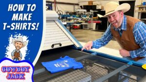Man wearing a cowboy hat and vest demonstrates screen printing a blue T-shirt on a large press in a workshop, for a tutorial video.