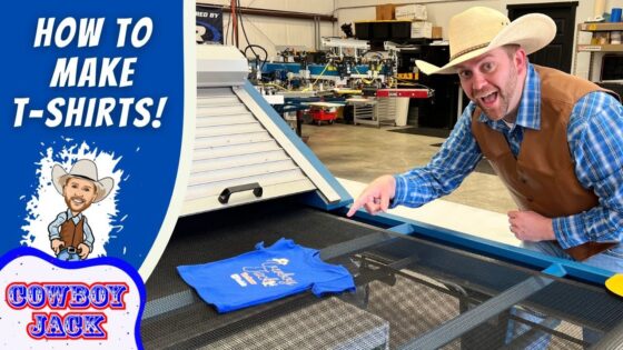 Man wearing a cowboy hat and vest demonstrates screen printing a blue T-shirt on a large press in a workshop, for a tutorial video.