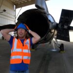 Maintenance worker in a high-visibility vest and hard hat poses with hands behind head beside a jet engine inside an aircraft hangar.