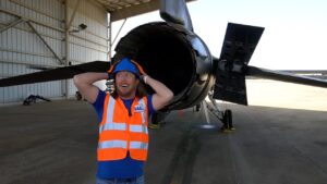 Maintenance worker in a high-visibility vest and hard hat poses with hands behind head beside a jet engine inside an aircraft hangar.