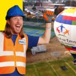 Cheerful construction worker in blue helmet and orange safety vest raises his arms beside a multicolored hot air balloon on a grassy field with a bright yellow background to the left.