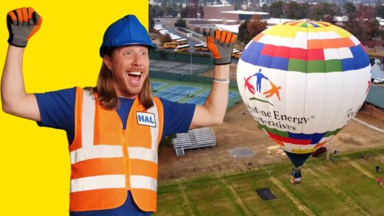 Cheerful construction worker in blue helmet and orange safety vest raises his arms beside a multicolored hot air balloon on a grassy field with a bright yellow background to the left.