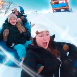 Kids sledding down a snowy hill on a blue inflatable tube, laughing and bundled in winter gear.