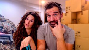 Two adults filming a video in a storage-like room with stacked cardboard boxes; the woman points at the camera while the man touches his eye with a playful, surprised expression on both faces.