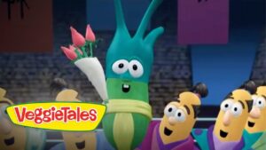 Green veggie character holding a bouquet of pink roses on stage, VeggieTales logo visible in foreground, smiling audience of veggies nearby.