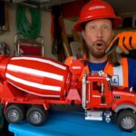 Man in safety gear (red hard hat, orange vest) holds a large red toy cement mixer truck in a cluttered garage.