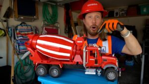 Man in safety gear (red hard hat, orange vest) holds a large red toy cement mixer truck in a cluttered garage.