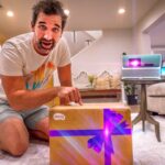 Man kneeling on a living room rug and unboxing a gift-wrapped box, with a small BenQ projector glowing purple on a nearby table.