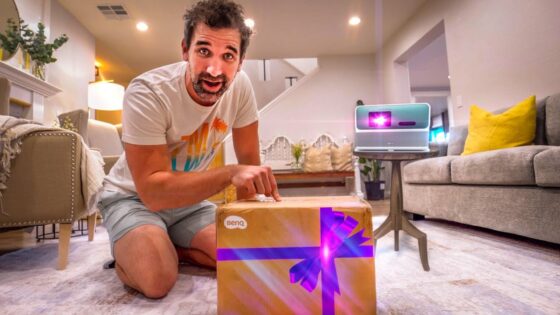 Man kneeling on a living room rug and unboxing a gift-wrapped box, with a small BenQ projector glowing purple on a nearby table.