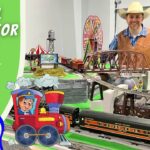 Smiling man in a cowboy hat beside a large model train setup for kids with a green banner reading 'Model Trains for Kids!'