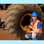 Mechanic in a blue hard hat and orange safety vest crouches beside a giant dusty tire, holding a power drill in a dusty yard workshop.