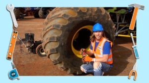 Mechanic in a blue hard hat and orange safety vest crouches beside a giant dusty tire, holding a power drill in a dusty yard workshop.
