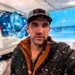 Man in a black and brown jacket and cap takes a selfie at a snowy loading dock with a blue truck behind him.