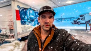 Man in a black and brown jacket and cap takes a selfie at a snowy loading dock with a blue truck behind him.