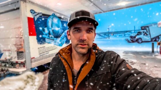 Man in a black and brown jacket and cap takes a selfie at a snowy loading dock with a blue truck behind him.