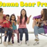 Group of people sitting on a bench: a guitar teacher or performer on the right, singing, with children and a woman, bright text 'I Wanna Bear Fruit' above.