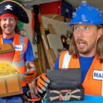 Two men in safety vests in a workshop, one wearing a pirate hat and the other a blue hard hat, with a cartoon treasure chest of gold coins in front.