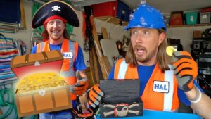 Two men in safety vests in a workshop, one wearing a pirate hat and the other a blue hard hat, with a cartoon treasure chest of gold coins in front.