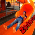 Man in a blue helmet and orange safety vest slides down a bright orange indoor tube slide, arms raised in excitement at a trampoline park, with a padded orange mat beneath.