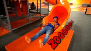 Man in a blue helmet and orange safety vest slides down a bright orange indoor tube slide, arms raised in excitement at a trampoline park, with a padded orange mat beneath.