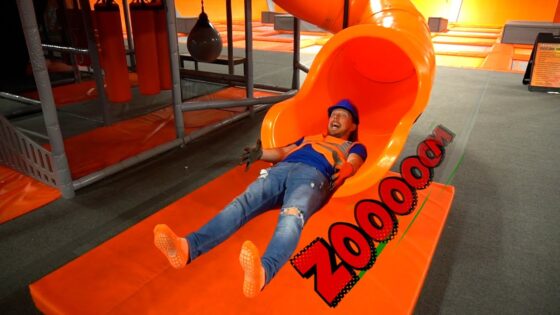 Man in a blue helmet and orange safety vest slides down a bright orange indoor tube slide, arms raised in excitement at a trampoline park, with a padded orange mat beneath.