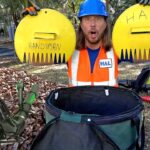 Man in a blue hard hat and orange safety vest holds two large yellow circular props labeled 'HANDYMAN' and 'HAL' outdoors, with yard tools and piles of leaves around.