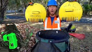 Man in a blue hard hat and orange safety vest holds two large yellow circular props labeled 'HANDYMAN' and 'HAL' outdoors, with yard tools and piles of leaves around.