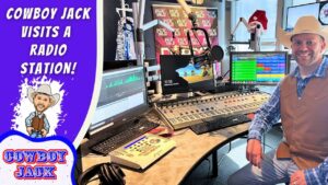 Smiling man in a cowboy hat and vest sits in a radio station studio with mixers and screens; banner reads "Cowboy Jack Visits a Radio Station".