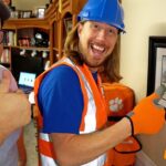 Two men smiling and giving thumbs up; one in a blue hard hat and orange safety vest, the other in a cap, in a home workshop backdrop with a framed painting and paw-print bag.