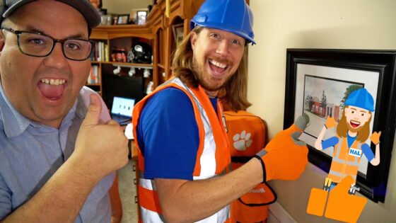 Two men smiling and giving thumbs up; one in a blue hard hat and orange safety vest, the other in a cap, in a home workshop backdrop with a framed painting and paw-print bag.