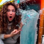Woman with a shocked expression as water bursts from a pipe in a DIY space, with yellow warning tape in the background on the wall behind her.
