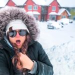 Person in a black parka with a fur-lined hood and sunglasses, taking a surprised selfie in a snow-covered street with colorful houses in the background, cars blanketed in snow beyond.