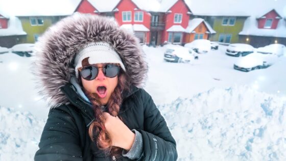 Person in a black parka with a fur-lined hood and sunglasses, taking a surprised selfie in a snow-covered street with colorful houses in the background, cars blanketed in snow beyond.