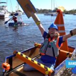 Woman in blue helmet and orange life vest cheers in a small DIY boat, cardboard wings raised above the water; harbor with boats and onlookers in the background.