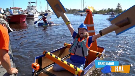 Woman in blue helmet and orange life vest cheers in a small DIY boat, cardboard wings raised above the water; harbor with boats and onlookers in the background.
